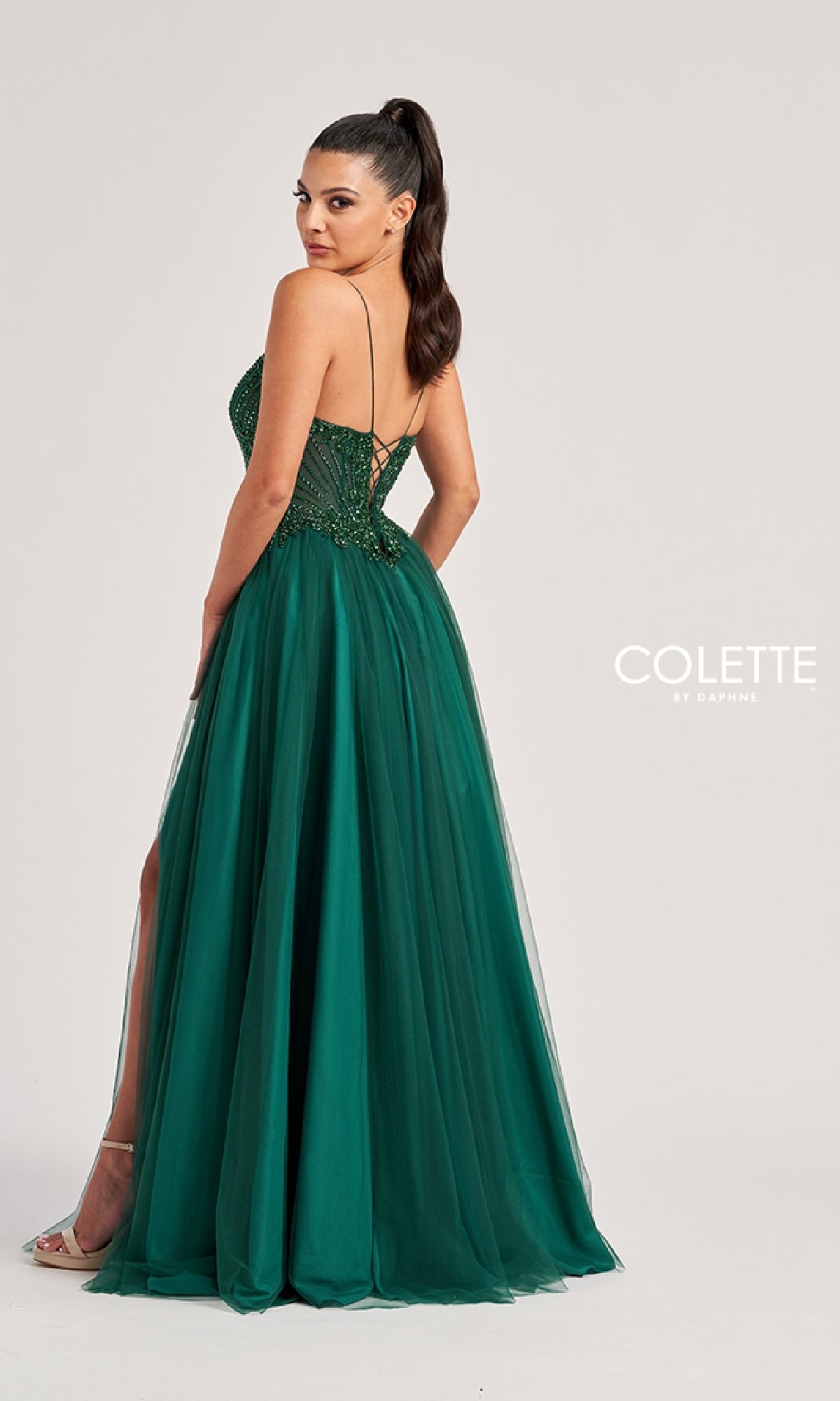 A woman in the Colette CL8250 long prom dress stands facing backward, glancing over her shoulder. The dark green gown features a strapless sequin bodice, tulle skirt with slit, and an elegant lace-up back.
